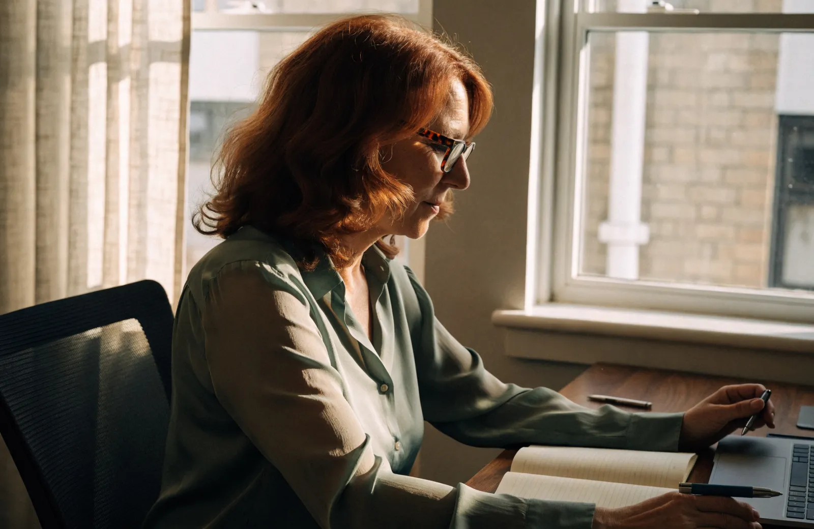 A red-haired woman in glasses writing in a notebook at a desk beside a sunny window
