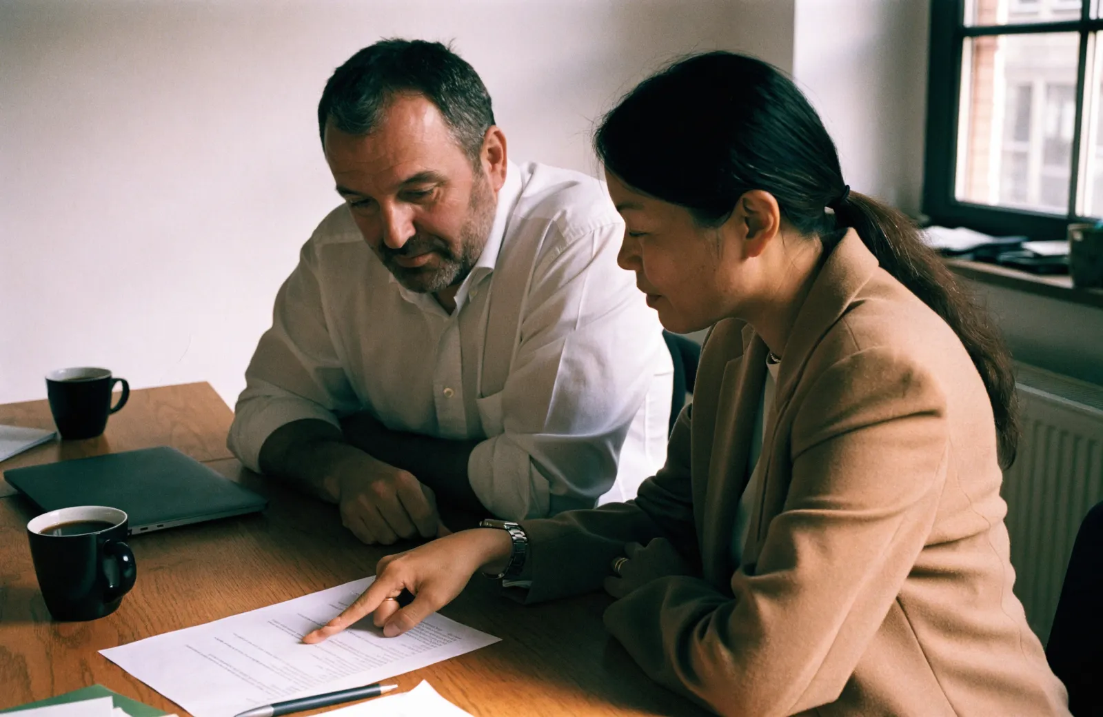A bearded man in a white shirt and a woman in a tan blazer reviewing a printed page at a wooden table