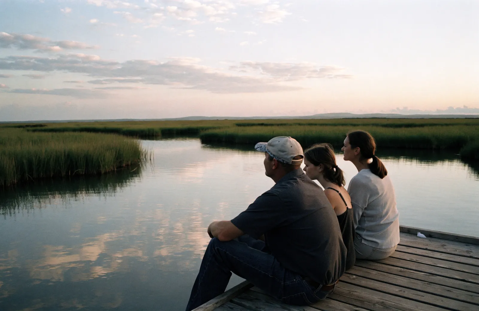 Three people seen from behind sitting on a wooden dock and looking out over calm marsh water at sunset