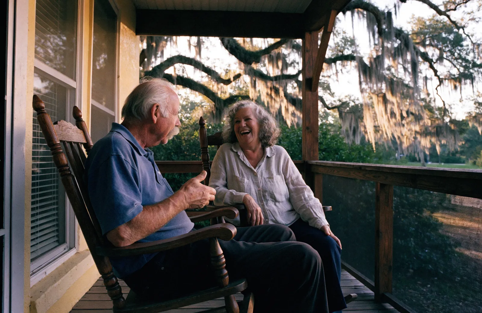 An older couple laughing in rocking chairs on a porch under live oak trees